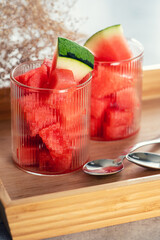 Two glasses filled with watermelon chunks and garnished with watermelon slices are placed on a wooden tray with a gray cloth and two spoons.