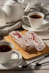 Two éclairs with white icing and red sprinkles on a wooden board, accompanied by a cup of tea and two spoons on a rustic wooden table.