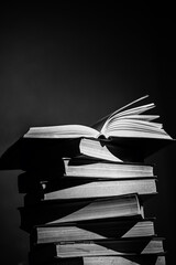 A stack of books with one open book on top, pages slightly fanned out, in a black and white setting...