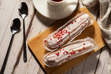 Two éclairs with white icing and red sprinkles on a wooden board, accompanied by a cup of tea and two spoons on a rustic wooden table.