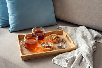 Two cups of tea on a saucer with two small tarts topped with white sprinkles, placed on a beige couch with blue pillows in the background