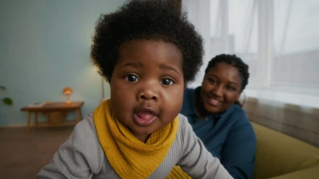 Portrait of curious African American toddler in yellow bib reaching for camera and pulling it closer, while her mother looking after her and smiling