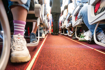 Low-angle view of airplane aisle with seated passengers and visible footwear during boarding,...