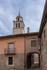 Fototapeta premium Historic bell tower rising above stone buildings in a quaint village, with a narrow arched passage and rustic charm under a partly cloudy sky