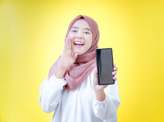 Naklejka premium Happy asian woman in a headscarf holds a smartphone, sharing excitement against a yellow backdrop.