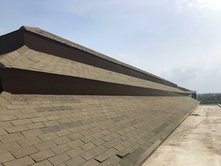 Weathered roof with asphalt shingles, partially lifted and deteriorated, captured from a low-angle perspective against a cloudy blue sky