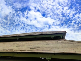 View of a worn asphalt shingle roof under a bright blue sky, showing visible damage and signs of aging on the roofing structure