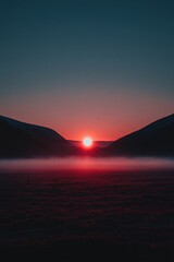 Red sun rising over misty field between dark mountains in a serene landscape at dusk