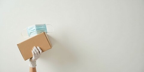 A gloved hand carefully holds a cardboard package with a protective face mask resting on top, signifying safe delivery and health consciousness.