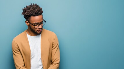 Fototapeta premium Young man with glasses and curly hair wearing a beige cardigan against a blue background with Copy Space