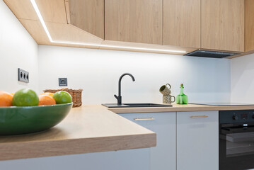 Small white kitchen with stylish, wooden cabinets, oven and sink with faucet. Bowl of fruits on the counter.  Bright interior of room in modern apartment.