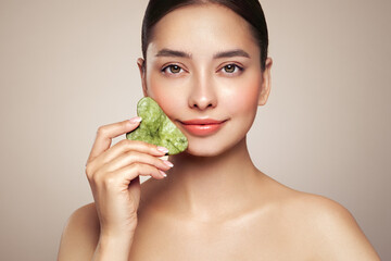 Beauty young woman doing skin care routine, scrapping, massage facial skin with jade gua sha...