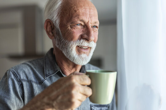 Cheerful senior man holding a cup of coffee and looking through the window at home. Pensive man looking through the window. - Powered by Adobe