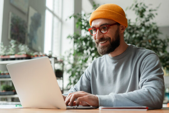 Smiling bearded man wearing glasses and beanie using laptop in bright office