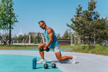 Fototapeta premium A muscular man in a blue tank top performs lunges with dumbbells in an open park area, demonstrating power and commitment in fitness.