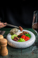 A close-up of a gourmet dish on a marble plate, featuring marinated beef, colorful vegetables, and sauces. A hand holds chopsticks, ready to savor the artfully presented meal.