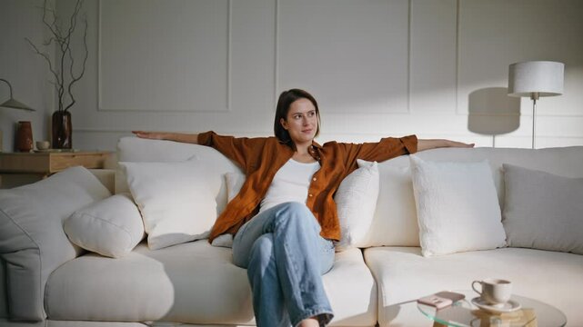 Happy woman lounging sofa modern living room. Relaxed brunette sit down couch 