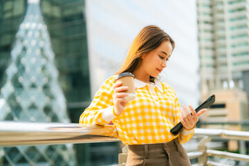 woman in yellow checkered shirt holding coffee cup and using tablet outdoors, with modern city...