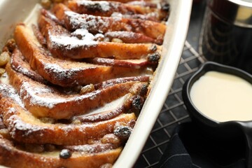 Freshly baked bread pudding in baking dish and custard cream on dark table, closeup