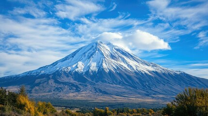 Majestic Snow Capped Mountain with Vibrant Blue Sky and Wispy Clouds in Clear Daylight, Surrounded by Lush Valley Landscape in Autumn.