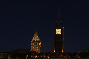 Fototapeta premium Night view of the illuminated Galata Tower and a modern clock tower in Istanbul. The warm lights contrast against the dark sky, creating a striking composition of historical architecture.
