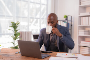 Naklejka premium Focused businessman is enjoying a cup of coffee while working on his laptop in a bright and modern office, surrounded by documents and charts, demonstrating a productive work ethic
