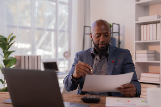 Concentrated african american entrepreneur examining paperwork and taking notes, working productively in a modern office environment, surrounded by laptop, charts, and bookshelves