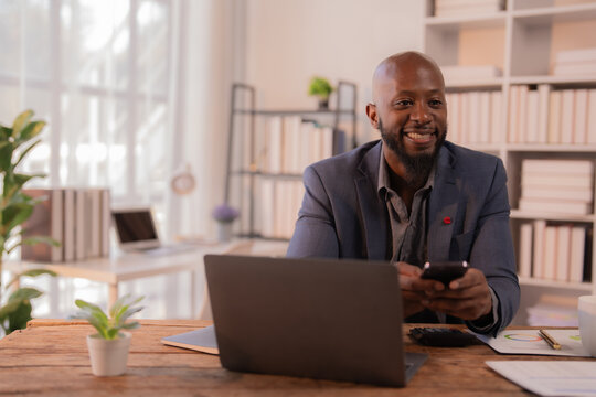 Happy african american manager is using mobile phone and working with laptop while sitting at wooden table in modern office with large window, bookshelves and plants