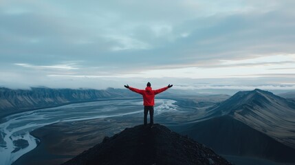 A figure in a red jacket stands triumphantly on a mountain peak, arms outstretched, overlooking a vast landscape of valleys and rivers under a cloudy sky.
