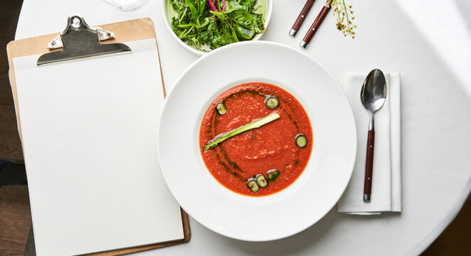 A bowl of rich tomato soup garnished with herbs sits on a dining table next to a fresh garden salad and a blank notepad, creating a cozy dining atmosphere