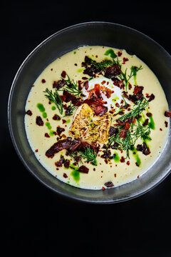 A rich, creamy soup is presented in a dark bowl, topped with crispy bits and fresh herbs, highlighting a delightful combination of flavors