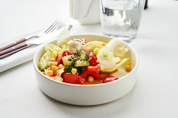 A bowl of fresh salad featuring a mix of lettuce, tomatoes, cucumbers, and chickpeas is placed on a dining table. A glass of clear water accompanies the dish, emphasizing a healthy meal choice