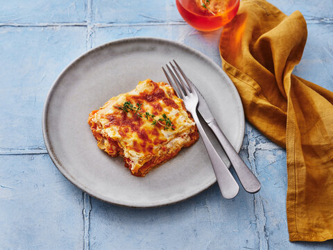 A rich lasagna is beautifully plated, garnished with fresh herbs, next to two silver forks. A refreshing drink sits nearby on a blue table covering, creating a inviting atmosphere