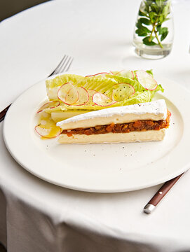 A beautifully plated gourmet dish includes layers of fresh vegetables, creamy sauce, and garnishes. The setting features a white tablecloth and a decorative vase, creating a refined atmosphere
