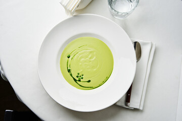 A bright green soup with unique swirls sits elegantly on a white plate, accompanied by a spoon and a glass of water, showcasing culinary artistry in a sophisticated setting