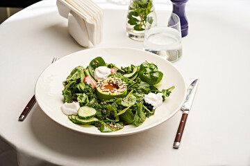 A vibrant green salad featuring fresh spinach, cucumbers, and avocado is elegantly presented on a white plate. Accompanying the salad is a light yogurt dressing, ideal for a healthy meal