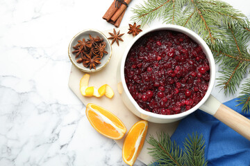 Tasty cranberry sauce in saucepan, spices, orange and fir branches on white marble table, flat lay. Space for text