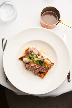 A succulent portion of beef rests on a delicate bed of roasted vegetables, elegantly plated at a sophisticated dining table. A drink is nearby, enhancing the gourmet experience