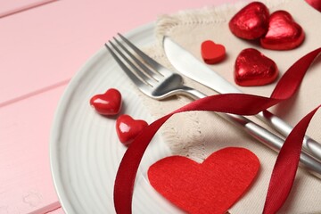 Romantic place setting for Valentine's day. Cutlery, napkin, plate and decorative hearts on pink wooden table, closeup