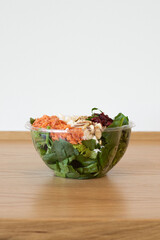 Fresh spinach salad with shredded carrots, feta cheese, cranberries, and sliced almonds in a clear plastic bowl on a wooden countertop