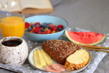 Tray with various healthy breakfast food, open book and reading glasses on the bed. Breakfast in bed concept. Selective focus.