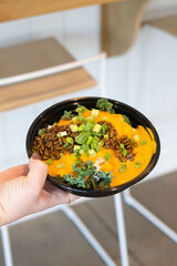 Hand holding a rice bowl topped with creamy orange sauce, roasted seeds, kale, and chopped green onions