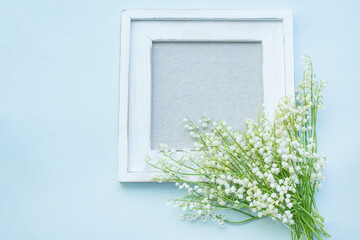A white wooden picture frame with a fabric-covered center, decorated with delicate  lily-of-the-valley flowers on a soft lavender background. top view. copy space 