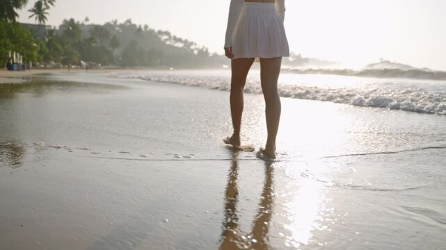 Female feet walking barefoot in sea surf waves. Person's legs in mini skirt going relaxed along sandy coast. Attractive woman strolling and enjoying sunset at the seaside on vacation. Low angle.