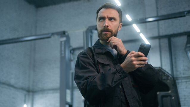 Mechanic man auto mechanical worker using mobile phone gadget in repair car service garage automobile factory pensive thoughtful male guy repairman look at smartphone screen think typing on cellphone