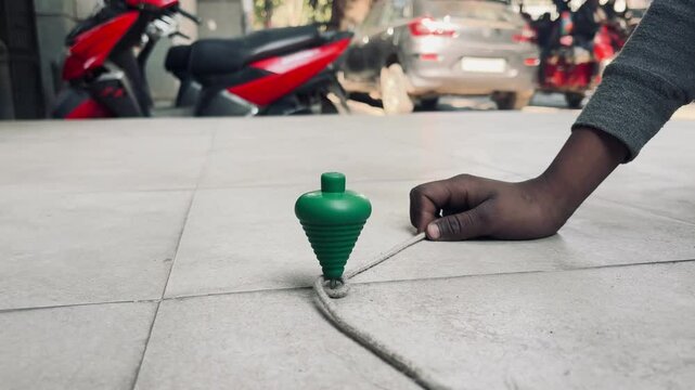 Spinning Top Fun: A child's hand skillfully winds the string around a vibrant green spinning top, preparing it for a playful spin on the pavement.