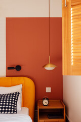 Close-up of wooden laconic classic bedside table with book and alarm clock. Cozy interior with bright accents. Bed with contrasting pillows.