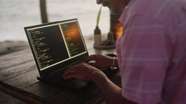 Web developer types code on laptop at beachside cafe with ocean view at dusk. Freelancer programs software, enjoys sea breeze, sunset vibes. Remote work, digital nomad lifestyle by the coast. Slowmo