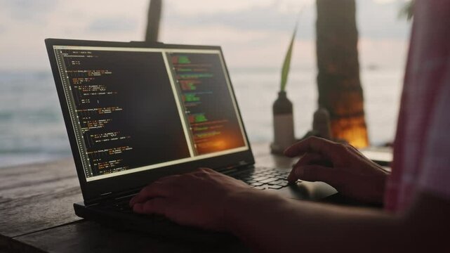 Web developer types on laptop at beach cafe with sunset over ocean. Male software coder creates code for project amid palms, sea. Remote work concept combines leisure, pro tasks. Slow motion.