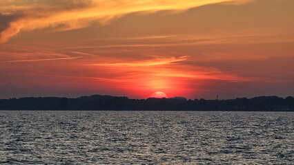 Atemberaubender Sonnenuntergang über einer ruhigen Wasserlandschaft. Der Himmel leuchtet in warmen Orange-, Rot- und Blautönen, während die Sonne am Horizont versinkt. Zarte Wolkenformationen und die 